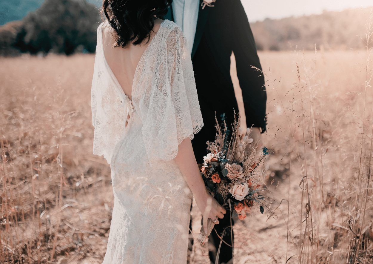 Braut im Spitzenkleid mit Strauß neben dem Bräutigam in einem goldenen Feld